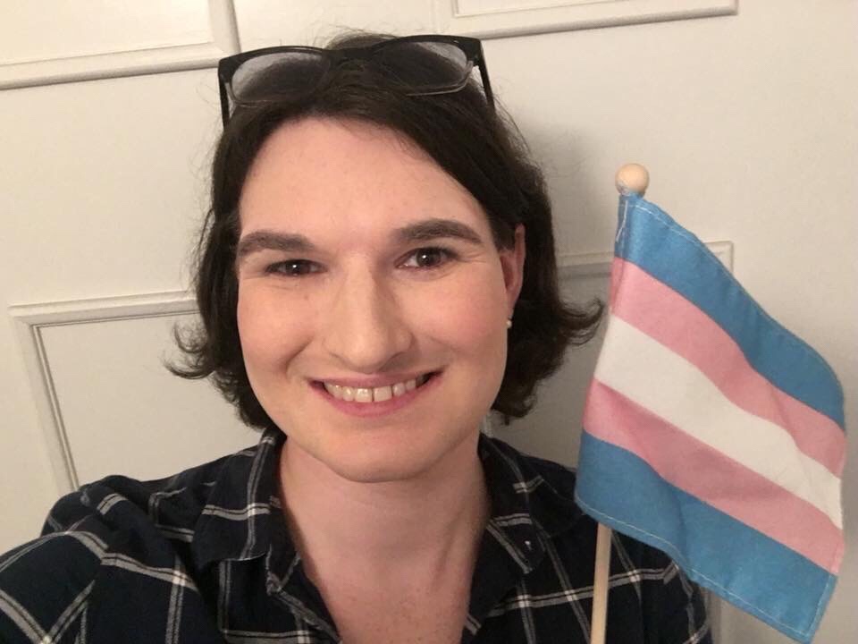 A woman with short dark hair is holding the Transgender Pride flag.