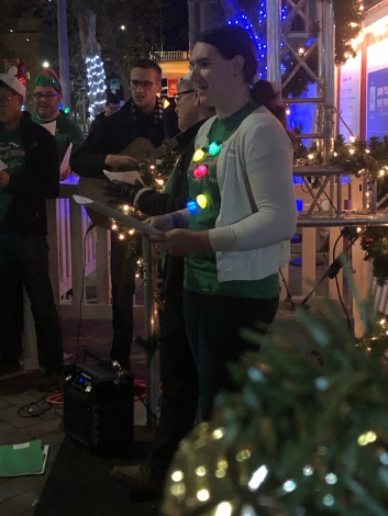 A woman stands in front of a group. She's wearing a green tshirt and holiday light necklace. She is singing.